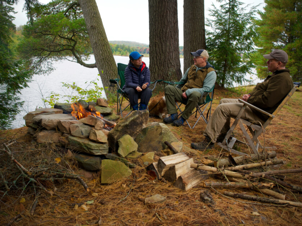 People sitting around a fire pit in the woods on a lake